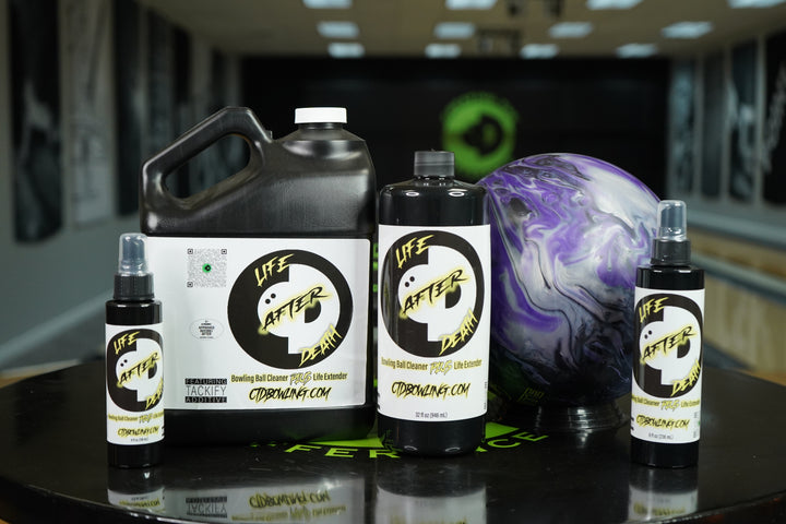 Various sized bottles of Life After Death Bowling Ball Cleaner + Life Extender on a table with a bowling ball in a bowling alley.